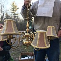 Chandelier held outdoors showing polished brass body, six arms with lampshades in beige/brown fabric with yellow trim.