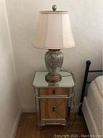 Mirrored side table with wooden drawer and cabinet topped with decorative table lamp against a beige wall and hardwood floor.