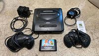 Overall view of Sega Genesis console with two controllers, AV cable, power adapter, and game cartridge laid out on floor.