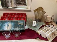 Showcase of the boxed Longchamp stem glasses, glass vase, bowl, candlesticks, and spiral candle boxes on a surface with burgundy cloth beneath.