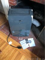 Front view of air conditioner with attached hose, power cord, remote and manual on floor