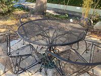 Top view of round wrought iron outdoor table with mesh top and radial decorative support design, chairs arranged around it outdoors on stone ground.