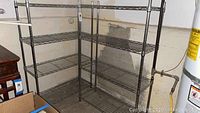 Photo showing two chrome metal wire shelving units set cornered against wall near water heater and wooden furniture, minor wear visible.