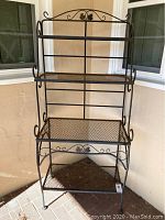 Full view of wrought iron bakers rack with four shelves and decorative scroll details on corners and legs. Shows rust spots and wear mainly on bottom shelves.