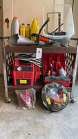 Photo showing red jump starter, various automotive fluids including antifreeze, funnel, tools, and jumper cables on and under a wheeled cart