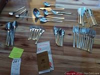 Full view of the flatware set arranged on wooden surface showing spoons, forks, knives, and serving utensils with detailed decorative handles, accompanied by certificates.