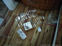 Full layout of mother of pearl handled flatware pieces arranged on wood floor with floral trivet in center.