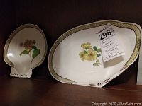 Two clam-shaped Wedgwood plates with floral designs and gold trim shown side by side on a shelf with a lot sticker attached to one plate.