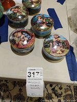 Photo showing five round porcelain Hummel music boxes with painted lids depicting children in pastoral scenes. All are trimmed in gold and arranged on a table.