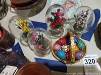 Four glass eggs on pedestal bases with flower arrangements inside and a basket filled with colorful small decorative eggs.