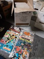 Box filled with multiple Star Trek comics in protective sleeves, some visible with vibrant covers showing sci-fi characters and title text