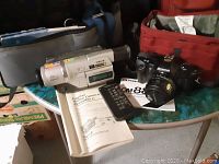 Sony DCR-TRV120 camcorder, remote control, Olympus OM-88 camera, cases and manuals on table.