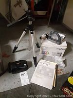 Photo of Hitachi camcorder on its box with manual and adjustable metal tripod standing behind it, additional black camera case on the floor