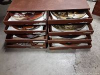 Eight collectors plates stored in flat wooden boxes arranged in two stacks of four with labels visible on drawer edges.