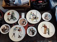 Eight collectors plates showing Norman Rockwell illustrations in various sizes, laid out on a black surface.