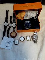 Multiple quartz battery-powered watches arranged on a surface with a MaxSold tag. Includes Stuhrling watch in orange box, several watches with leather and metal bands, and some watches without bands.