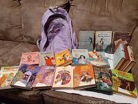 Photo showing 18 youth books of various titles laid out in front of a purple Roots backpack on a couch. Books include Nancy Drew numbers 8, 24, 41; three Donna Parker books; multiple Trixie Beldon books and other similar vintage youth books.