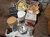 Photo showing an arrangement of kitchen items on a brown couch including a white coffee maker, shaped Wilton cake pans, bamboo steamer, wooden salt and pepper shakers, handheld grater, and various cake pans.