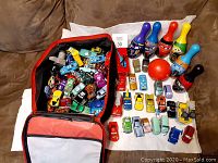 Open Lightning McQueen suitcase with over 70 toy cars, bowling pins, bowling ball, and additional Cars vehicle toys displayed on a white cloth on a couch