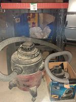 Larger red and black Shop Vac wet/dry vacuum with hose attached and smaller 12-volt wet/dry vacuum on floor.