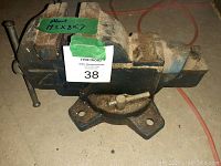 Side view showing the large metal vise with swivel base mounted on concrete floor. Green tape with handwritten dimensions approx 11.5 x 8 x 7 inches.