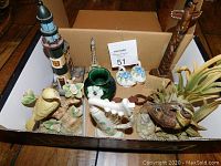 Nine assorted knick knacks in a box including music box with flowers, Royal Adderley swan salt and pepper shakers, Irish harp, green signed vase, Meadowlark bird figurine, Welsh lovespoon, Boma totem pole, wooden lighthouse, and metal Eiffel Tower.