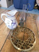 Photo showing a set of three antique items including a brass footed round trivet with intricate cut-out floral design, a purple and white patterned ceramic pitcher, and a brass or bronze wall candle holder with a glass chimney and loop handle.