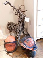 Photo showing two orange Stihl helmets with wire visors and padded ear protectors placed on floor next to a woven reindeer decoration made from natural twig materials.