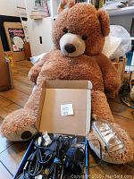 Large brown stuffed teddy bear seated on wooden floor with box of assorted wires and computer mouse in front.