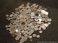 Photo of a large pile of assorted world coins on a dark surface showing variation in size and color.