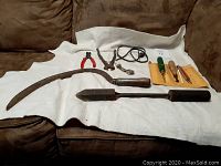 Full lot displayed on white cloth, showing antique curved scythe, blacksmith tool with copper end, wire cutters, pliers, and a set of small tools in a yellow pouch.