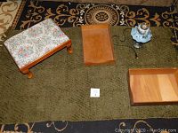 Lot contents showing footstool with needlepoint top, two wooden box trays, and metal desk lamp with glass shade on green rug placed over a gold and black patterned rug.