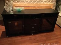 Full front view of black media cabinet showing top surface, three open cubbies, two drawers, and two glass-front cabinets