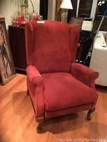 Front view of burnt red wing back recliner with curved wooden legs and rolled arms on hardwood floor.