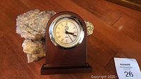 Small quartz mantle clock set in dark wood base with rounded top and silver bezel, placed on wooden floor next to rocks.