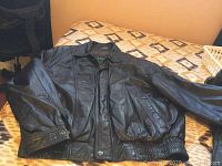 Black leather bomber jacket laid flat on bed showing front with zip and snap button closure and side pockets.