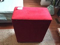Side view of the red velour-covered hamper sitting on hardwood floor next to a rug and bed.