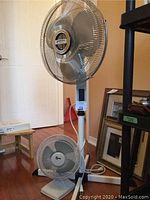 Two fans shown together: taller white floor standing fan and smaller white tabletop fan on floor near baseboard and framed artworks.