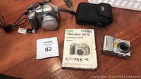 Canon PowerShot S2 IS camera, USB cord, instruction manual, Pantex OPTi camera, and seller tag on wooden floor