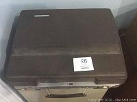 Closed hard brown plastic travel case for the typewriter, rectangular with handle on front.