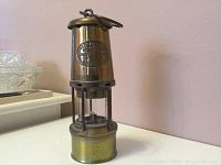 Full view of brass vintage miners safety lamp standing on a white surface in front of pink wall.