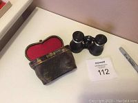 Mini black antique binoculars and worn leather carrying case with red felt lining.
