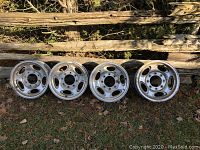 Photo of four aluminum car rims leaning against rustic wooden fence outdoors, showing wear and dirt.