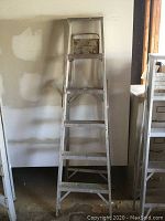 Front view of a 6 foot aluminum step ladder against a wall, showing all six steps and top platform, with visible wear and paint marks.