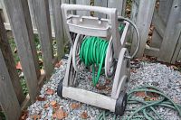 Front angle view of beige plastic hose reel cart with green garden hose on it, near wooden fence, gravel ground.
