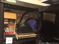 Photo showing a boxed Coleman lantern, black BBQ grill pan, and a black manual air pump on a shelf along with a sleeping bag in a black storage bag and a blue sleeping bag in a blue storage bag.