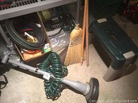 Photo showing Earthwise electric corded weed trimmer with coil green garden hose attached, black garden hose coiled on the floor, corn broom and two extra wooden handles leaning against wall, small orange gardening tray on floor.