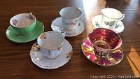 Five sets of tea cups and saucers arranged on wooden table, showing different colors and patterns