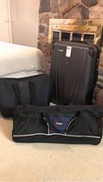 Photo shows three bags next to bed: one large black hard sided suitcase and two black soft sided bags.