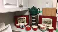Image showing green teapot atop boxed stack of four white rice bowls with chopsticks and six red mini bowls, Abbott mugs, and wooden placemats with UK scenes in background.